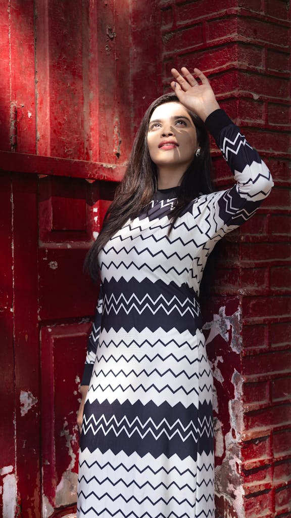 Fashionable woman in zigzag dress poses against red brick wall, looking upward.