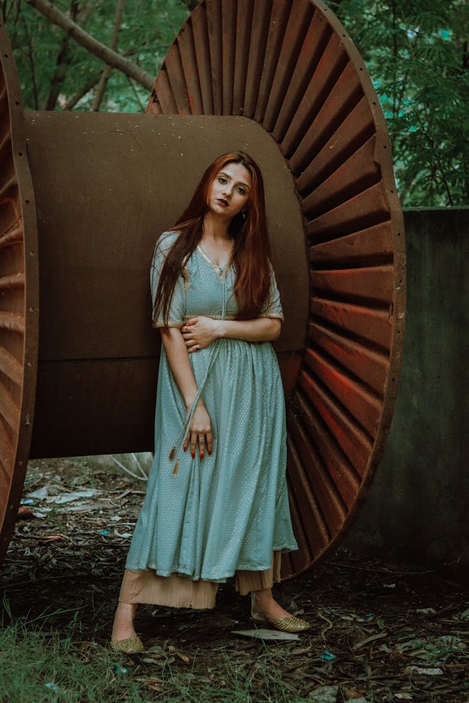 Full body of young romantic lady with long foxy hair in maxi dress standing near rusty metal coil in forest and looking at camera