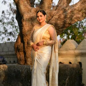 A confident woman in a traditional saree poses gracefully by a large tree.