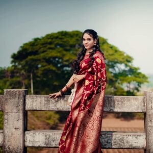 A woman wearing a red ornate saree stands outdoors by a fence, embracing cultural elegance.